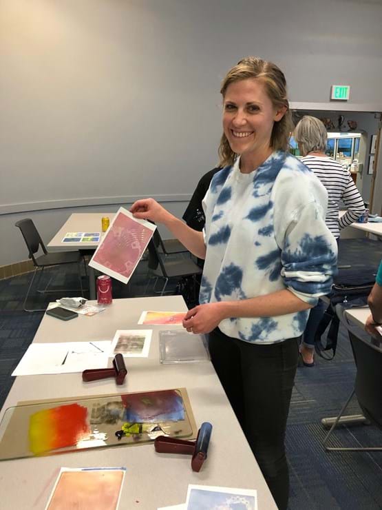 Woman wearing white and blue tie die shirt, holding up paper made art, smiling at the camera