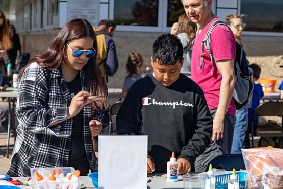 A boy wearing a black sweatshirt, and a girl wearing a plaid shirt and sunglasses looking down at wood rounds, making crafts at a table with people in the background