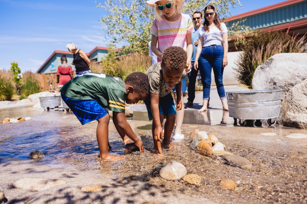 Children collect large stones from ankle-deep water in The WaterShed's simulated river as parents observe from nearby.