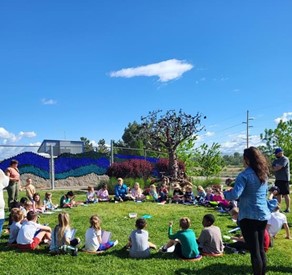 Several children sit in the grass in a large circle while volunteers lead them in an outdoor group activity.
