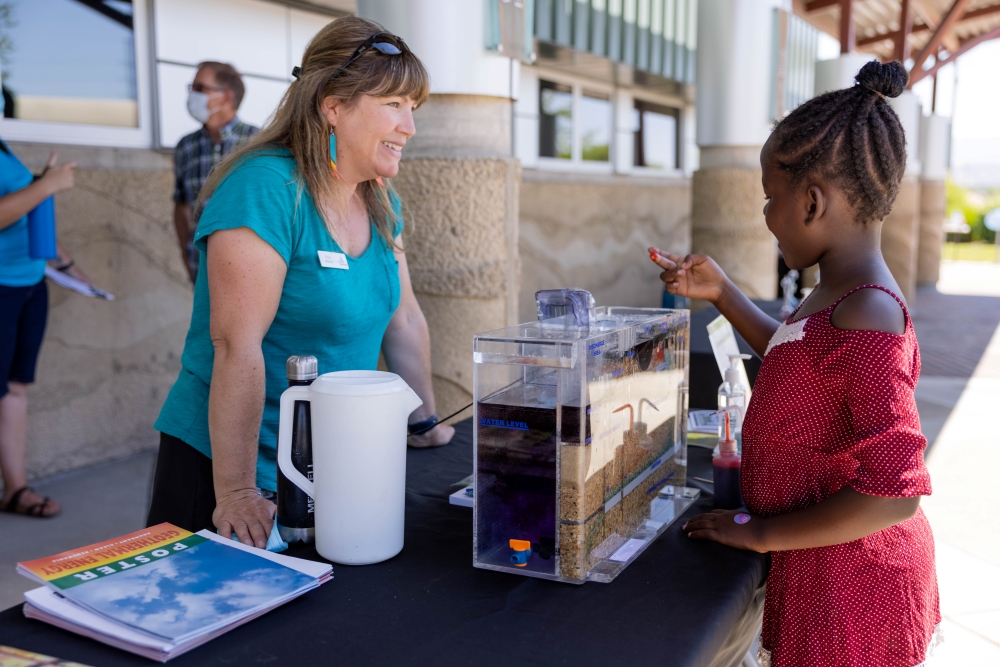 A young visitor and staff member discussing a demonstration model during an outdoor event at The WaterShed.