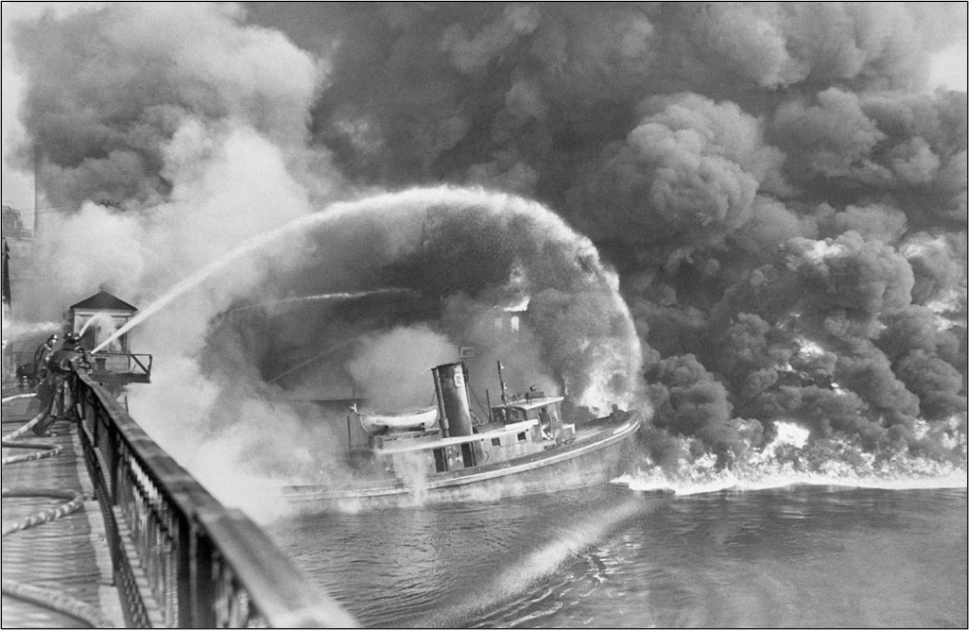 A boat is surrounded by a burning river, and firefighters are on a bridge spraying the boat with a hose from a distance.