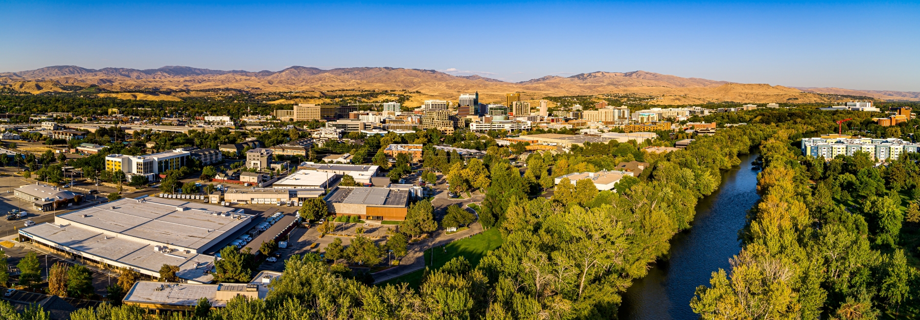 A panoramic aerial view of Boise, focused on the Boise River and surrounding greenery, with the downtown skyline in the background.